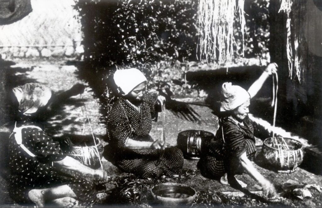 Black-and-white photo of three women in headscarves seated outdoors working fibre into thread beside baskets and bowls, with long strands hanging above