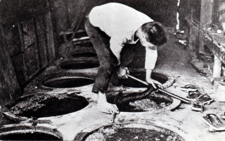 Black-and-white photo of a barefoot man bending over a row of round indigo vats set into the floor, stirring one with a long tool