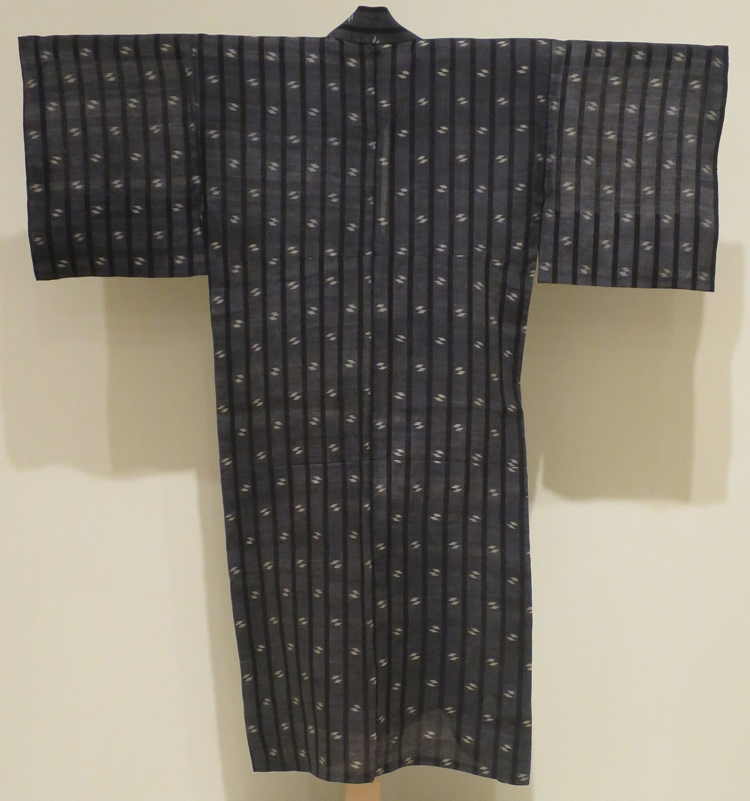 Dark blue-grey kimono hung on a wall, with rows of small white kasuri marks and fine vertical stripes