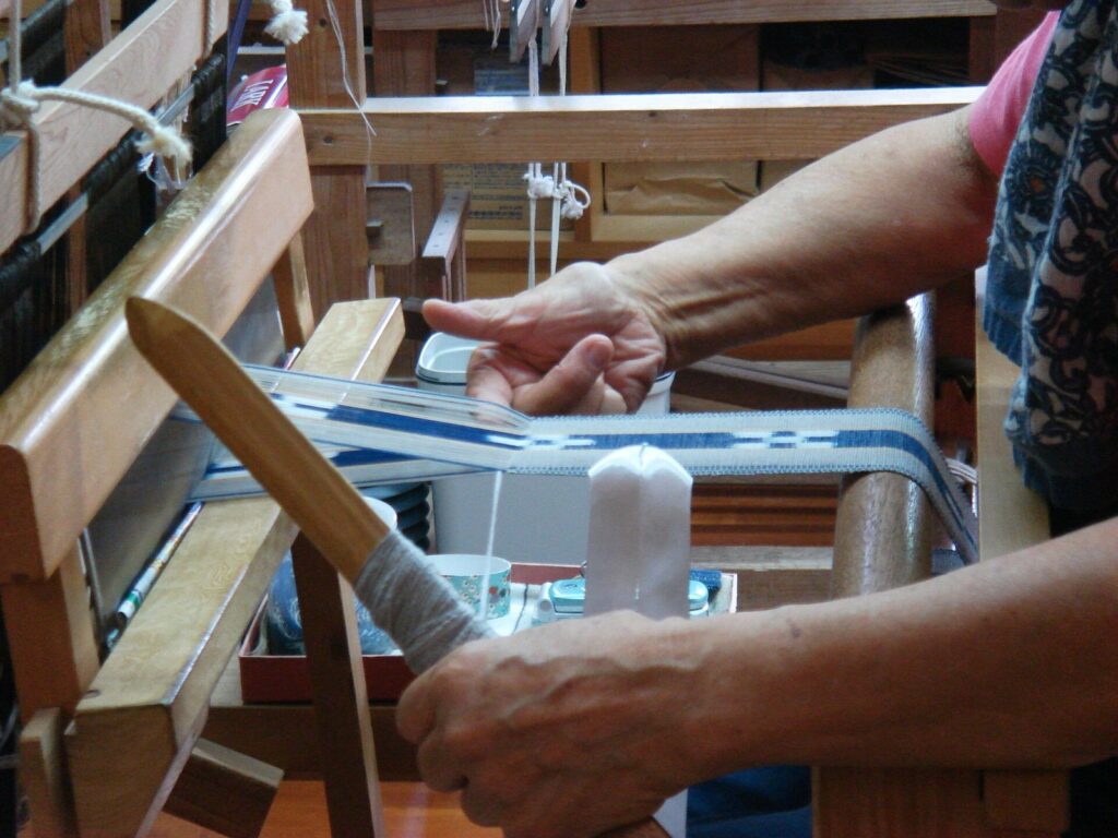 Close-up of hands at a wooden loom weaving a narrow blue and white minsā belt, with a shuttle wound with white thread
