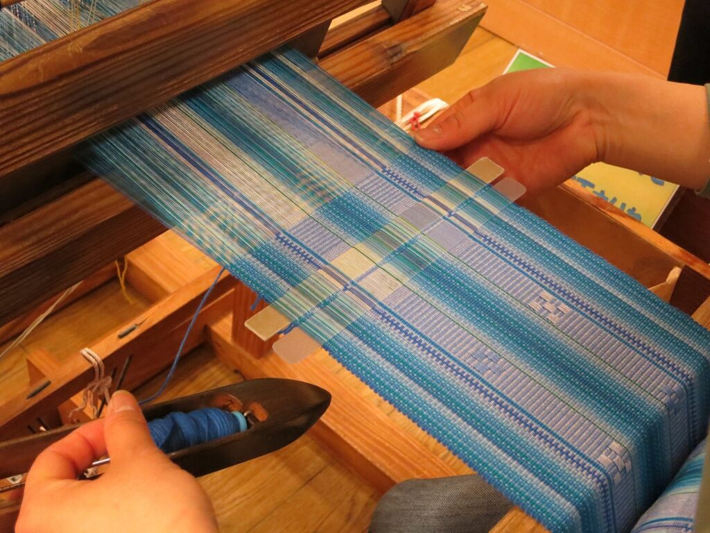 Hands weaving blue minsā cloth with the five-and-four pattern on a wooden loom in Ishigaki