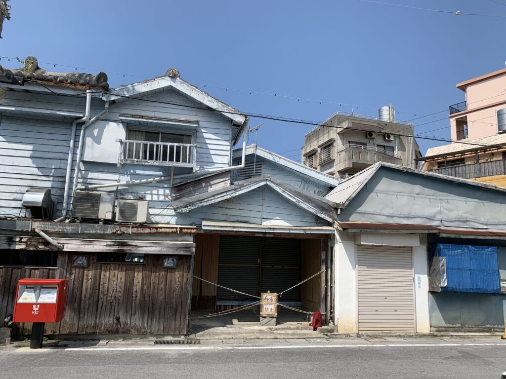 Weathered old wooden buildings with a red postbox in front, on a street in Naha