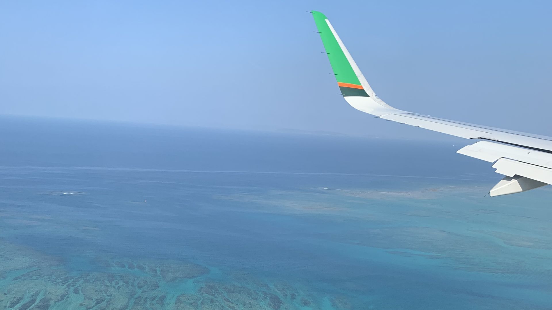 Aircraft wingtip over turquoise reef waters and the open sea, seen from a plane window flying into Okinawa