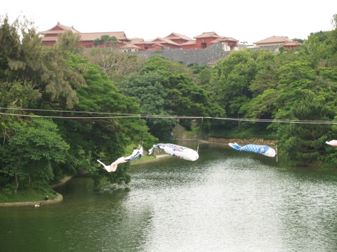 A green pond surrounded by trees with carp streamers strung across it and red-roofed castle buildings on the hill above