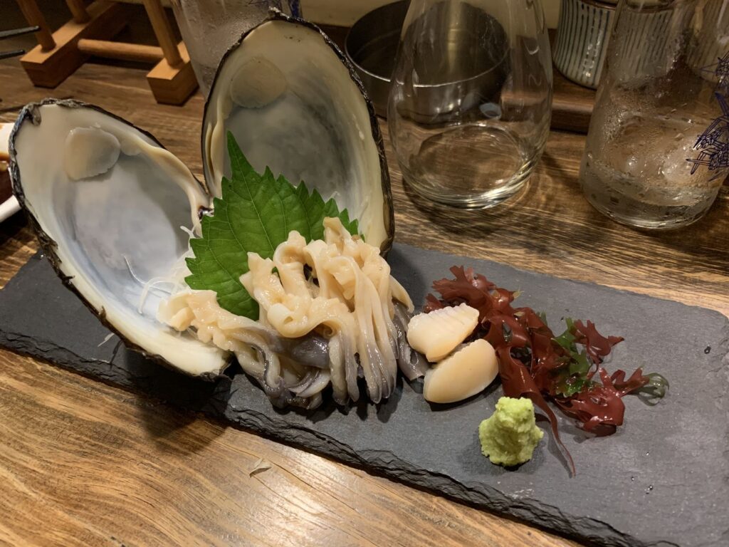 Sliced raw shellfish served in its shell with a shiso leaf, wasabi and garnishes on a slate plate