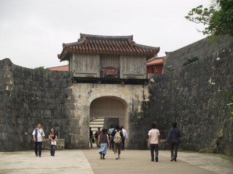 Visitors walking toward an arched stone gate with a tiled-roof tower set in Shuri Castle's stone walls