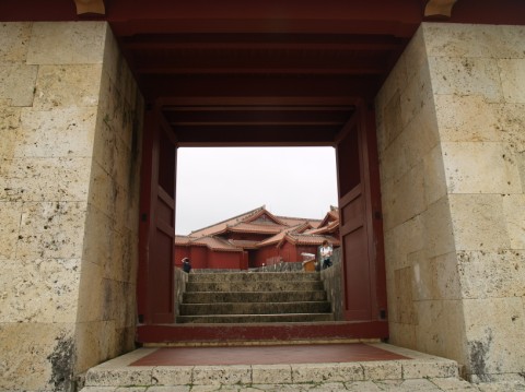 View through a red-painted gate in thick limestone walls to steps and a red building beyond, Shuri Castle