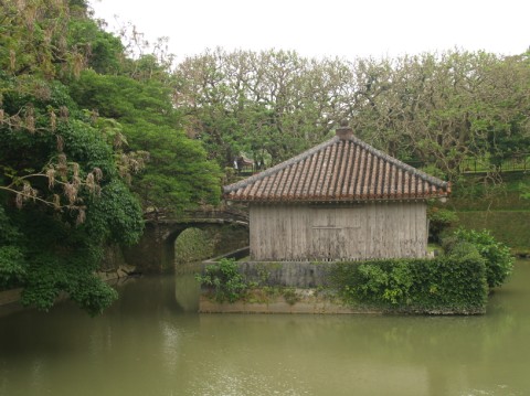 A small wooden hall with a tiled roof standing on an island in a pond, reached by a stone bridge, in Shuri