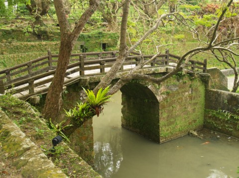 An old moss-covered stone arch bridge over a pond, framed by trees, in Shuri