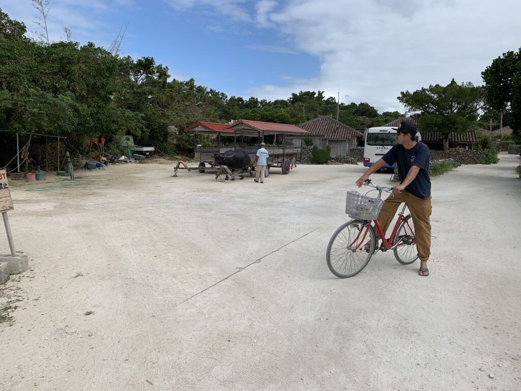 A cyclist riding along a wide white sand road with red-tiled houses behind, Taketomi