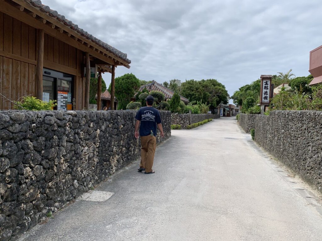 A white sand lane between coral-stone walls and a wooden house, with a person walking in the distance, Taketomi