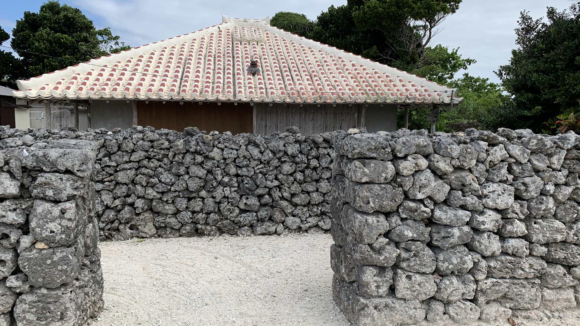 A traditional red-tiled house behind a coral-stone wall with an opening onto a white sand yard, Taketomi